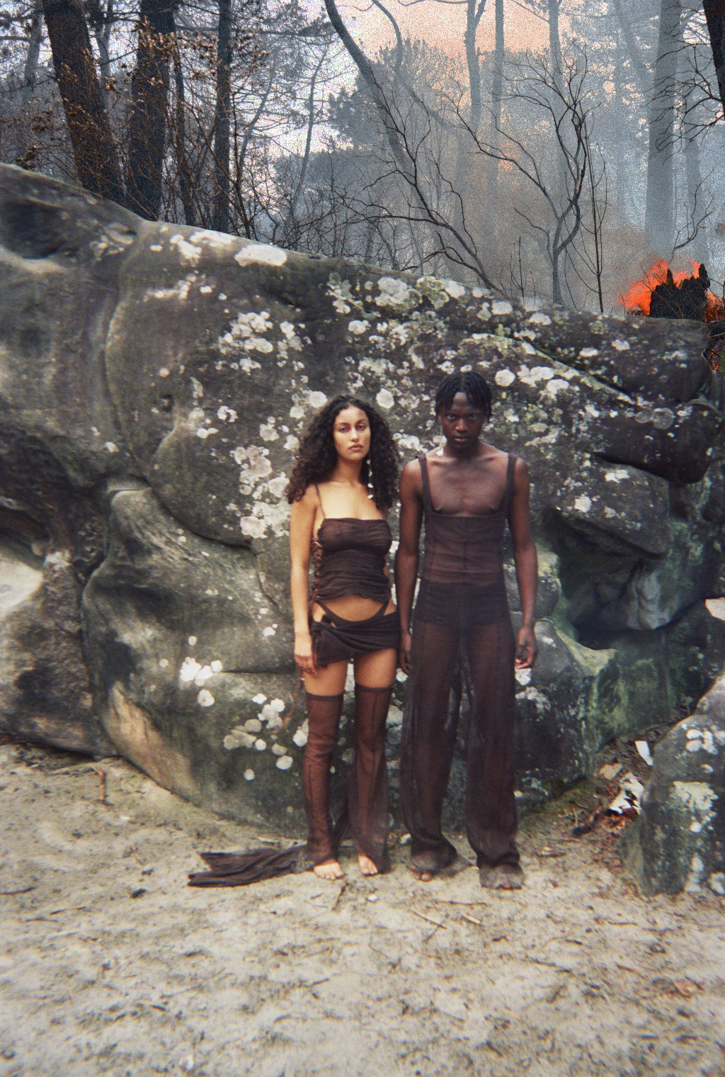 Une homme et une femme se tiennent devant un rocher, sur un sol sableux. La forêt derrière est sombre, et une flamme apparait.